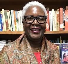 Ekua, an African-American woman with short, white hair, glasses and a nose ring stands, smiling in front of a bookshelf.